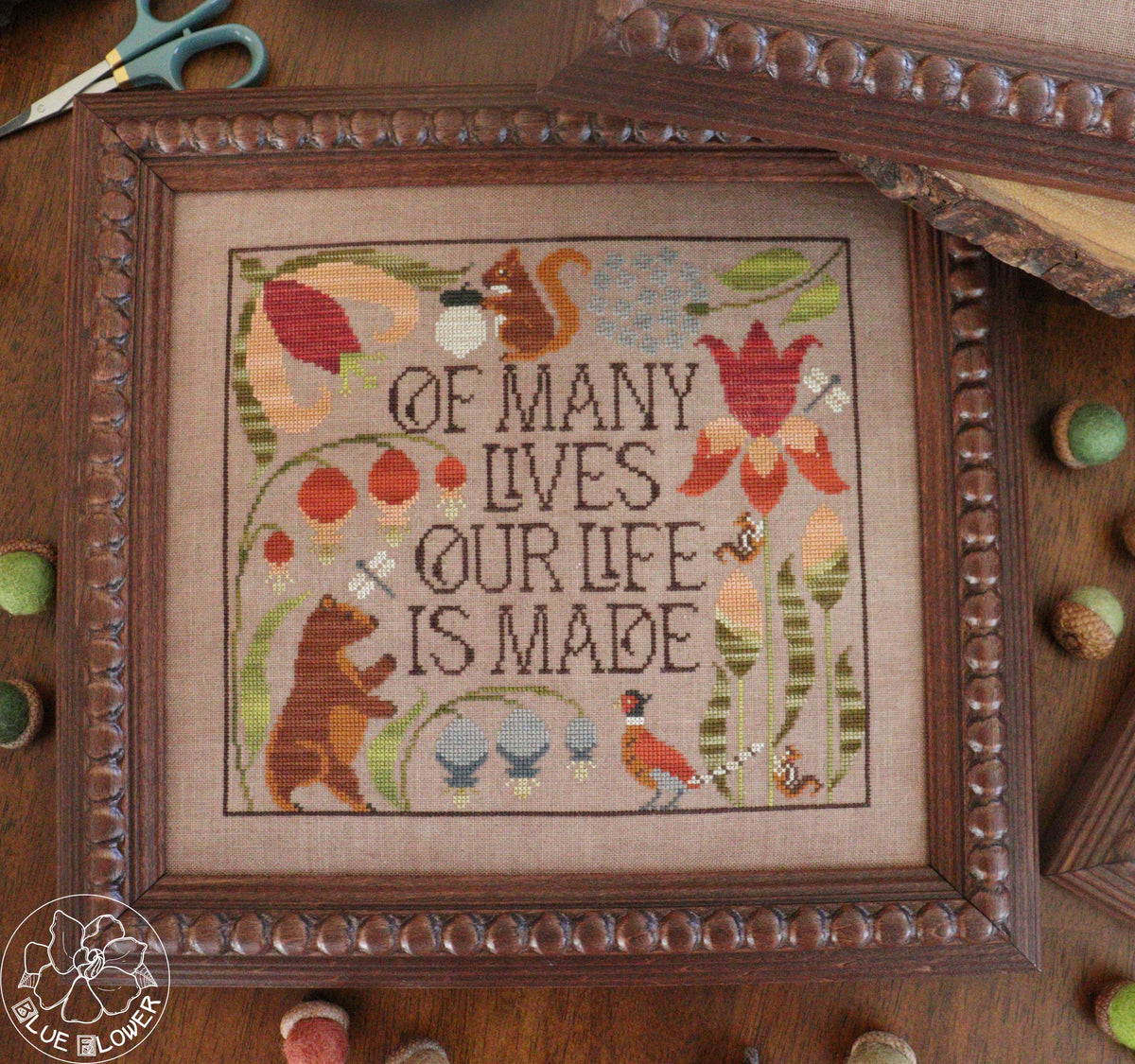 framed cross-stitch artwork featuring the phrase “OF MANY LIVES OUR LIFE IS MADE” surrounded by embroidered animals, flowers, and foliage