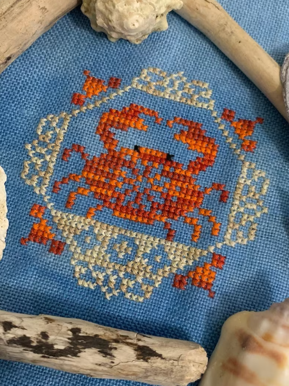 Cross stitch kit related to the Quirky Quaker Crab design, featuring a blue fabric with an embroidered crab and beach accoutrements.