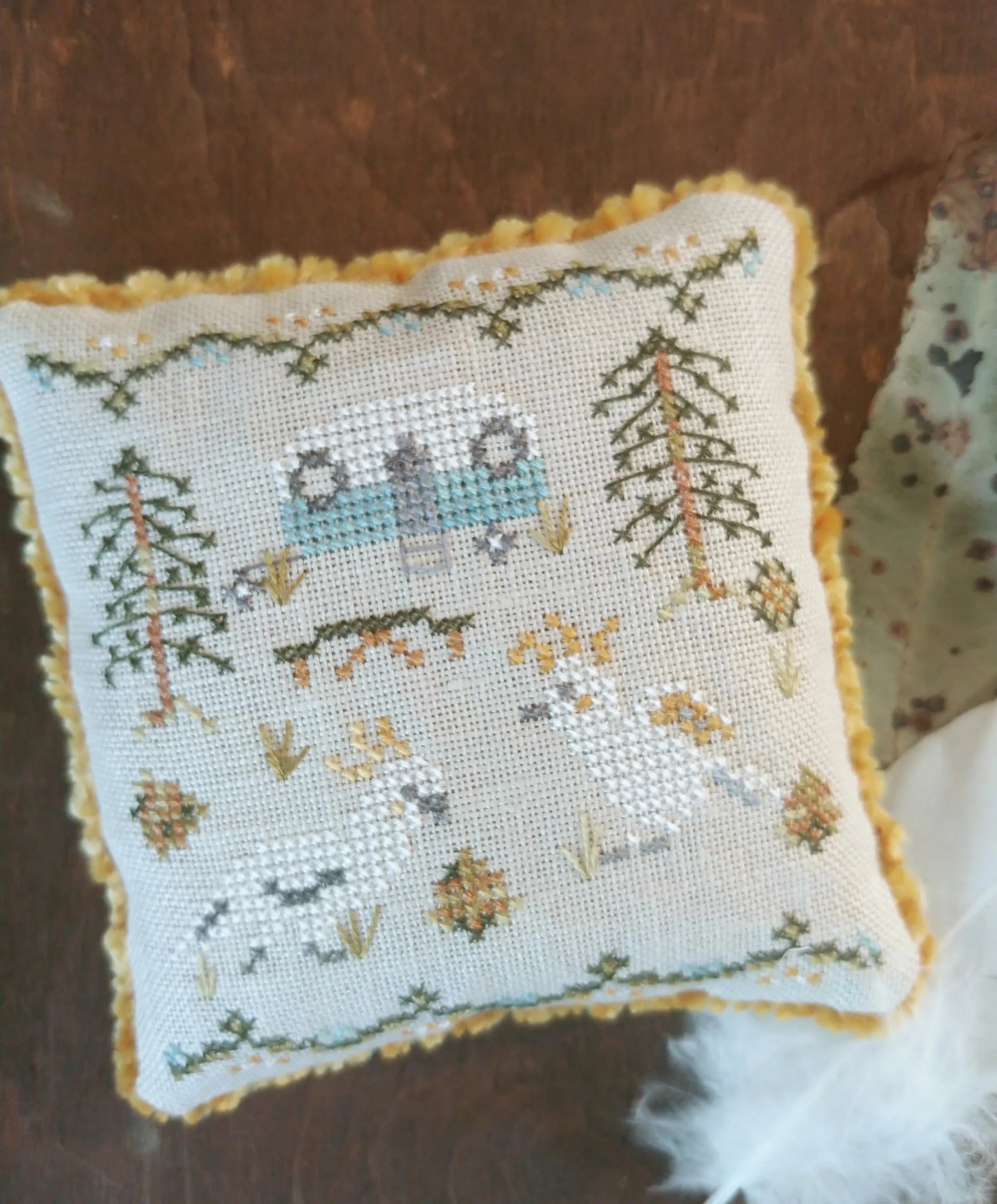 Decorative pillow with cross-stitched design of cockatoos and a caravan in Australian bushland on a wooden surface.