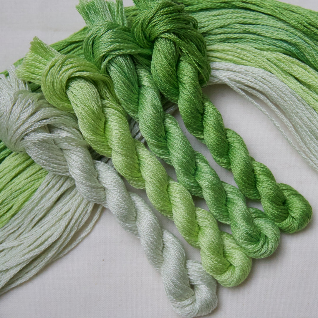 Four twisted skeins of embroidery thread in shades of grassy green through to pale mint, arranged on a light fabric background.