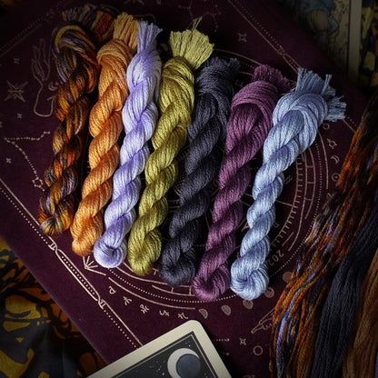 Seven twisted skeins of embroidery floss in assorted colours. The colours include orange-brown, lavender, mustard yellow, charcoal grey, mauve purple, and pale blue.