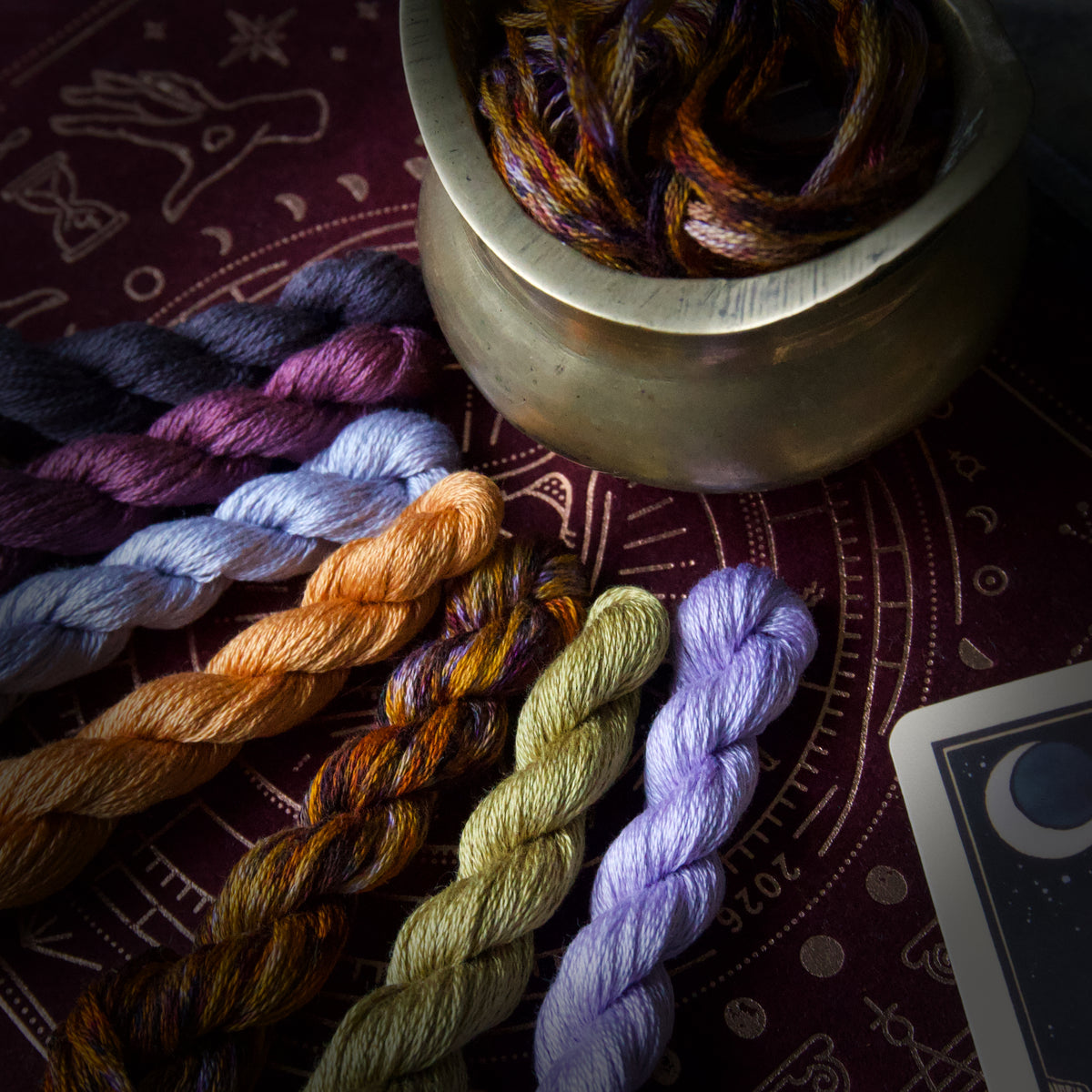 Six twisted skeins of colourful embroidery thread. The thread colours include purple, light blue, orange, brown, olive green, and lavender.