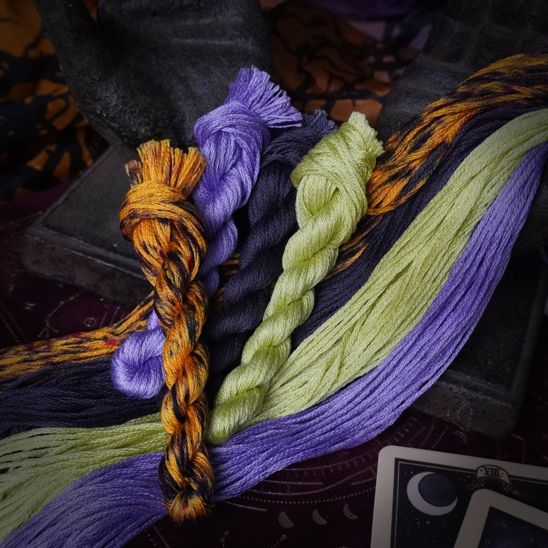 Four twisted strands of embroidery floss in purple, lime green, orange, and a black-and-orange pattern.