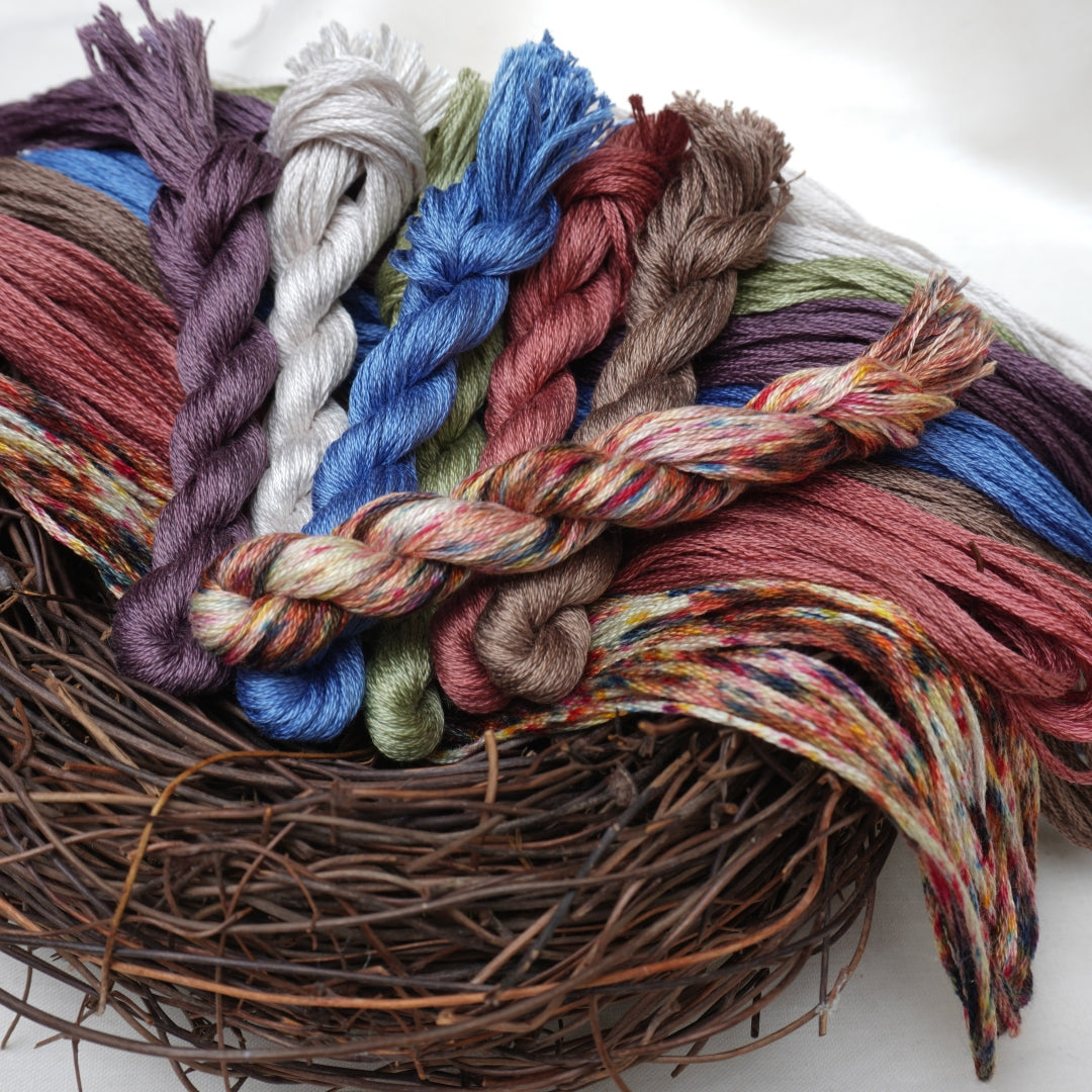 A close-up of multiple twisted skeins of embroidery thread arranged on a rustic woven nest base against a soft white background.