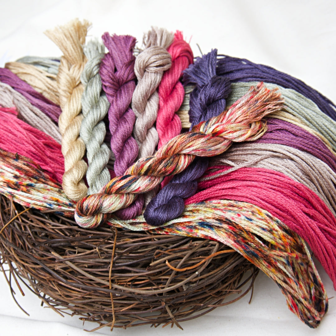 A close-up of variegated embroidery skeins in bright autumn colours arranged on a rustic nest base against a white background.