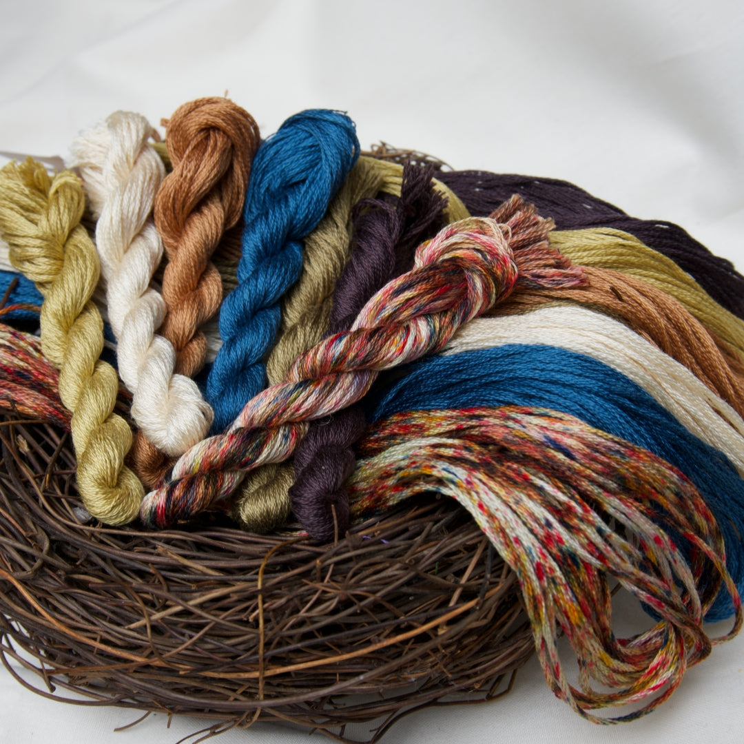 A collection of colourful twisted embroidery thread skeins in muted autumn tones, arranged on a rustic brown nest against a clean white background.