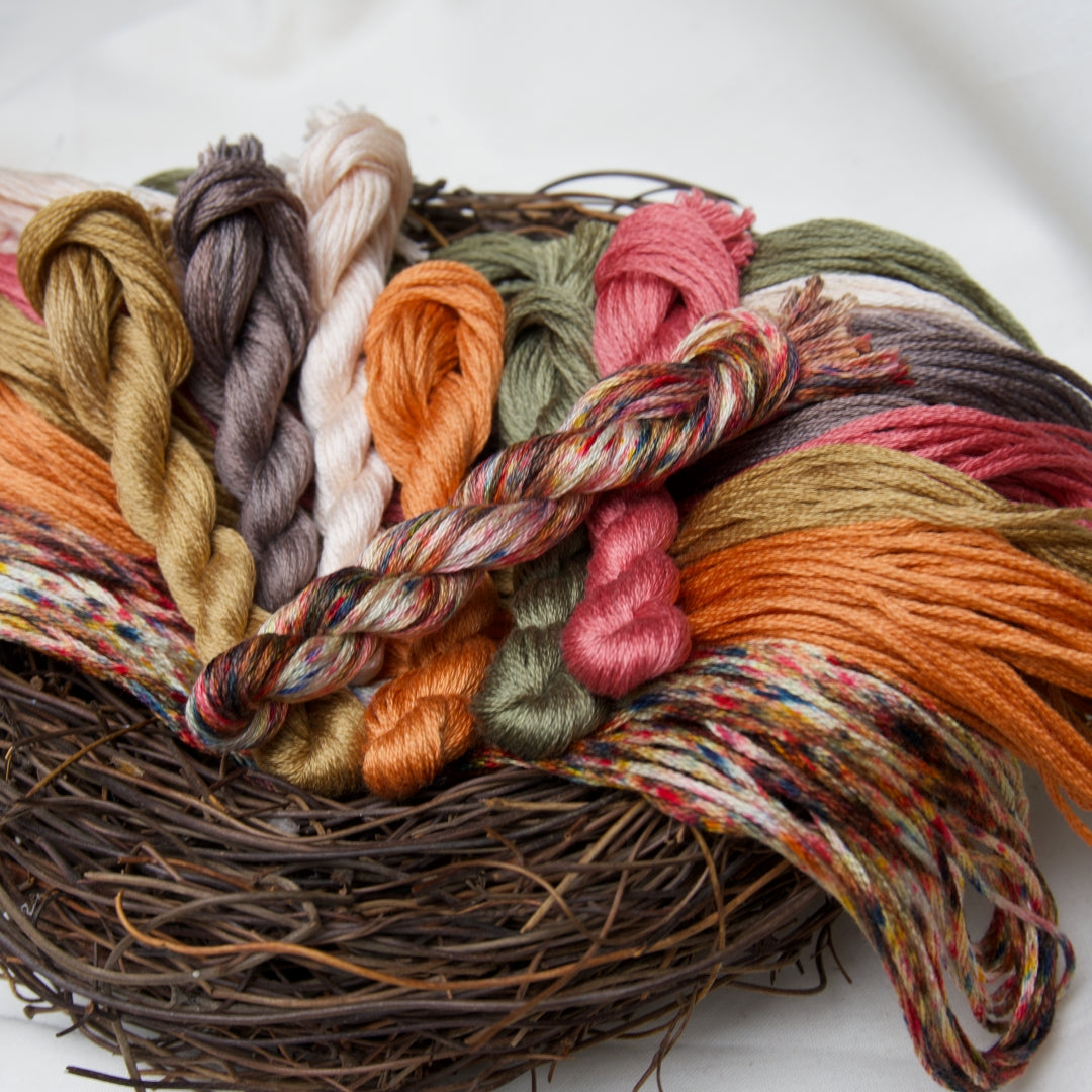A close-up of multiple skeins of twisted embroidery floss in autumn harvest colours arranged on a rustic woven nest base against a soft white background.