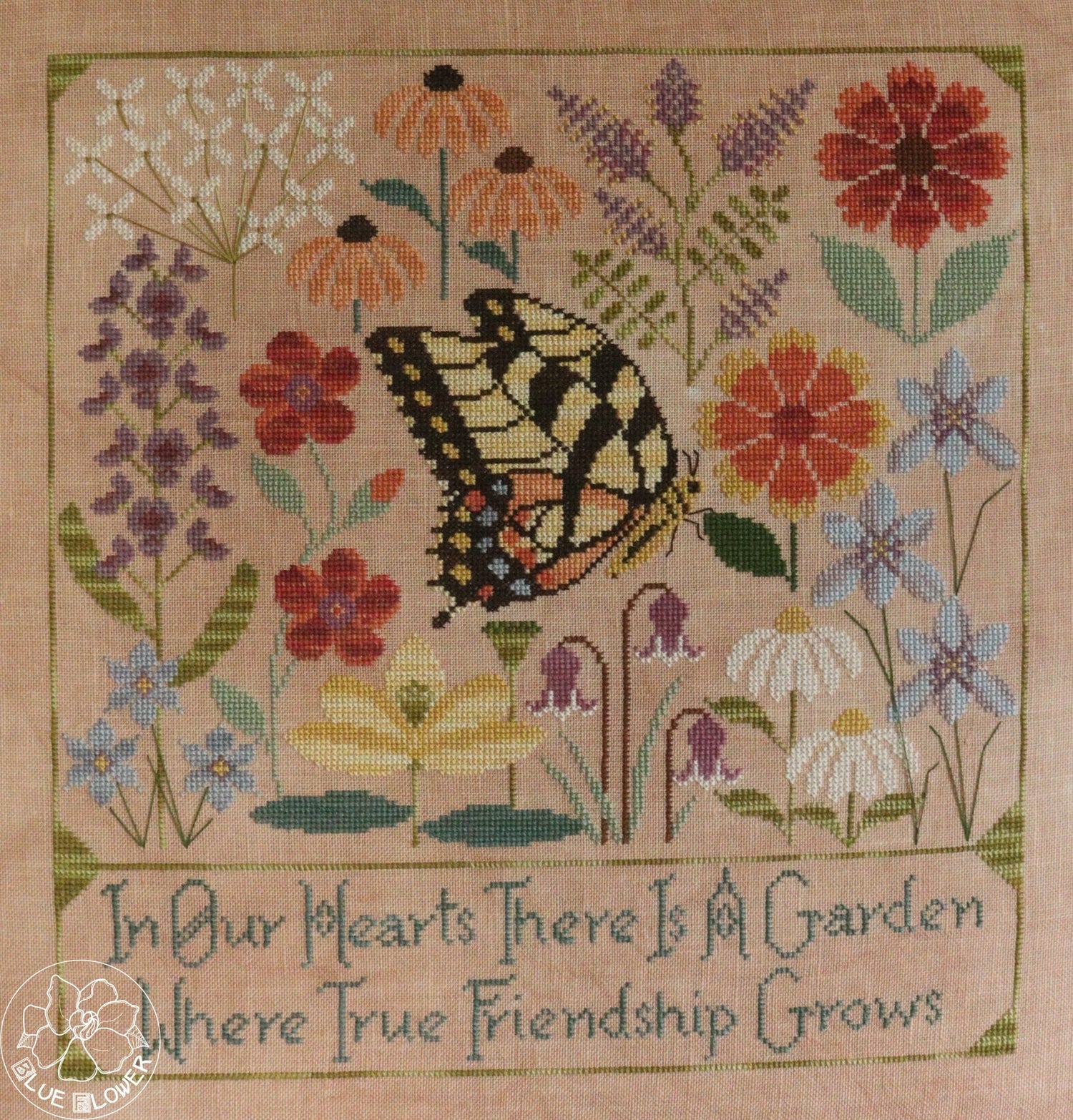 A square cross-stitch sampler featuring a yellow and black swallowtail butterfly surrounded by colorful wildflowers on a beige fabric background, with the embroidered text &