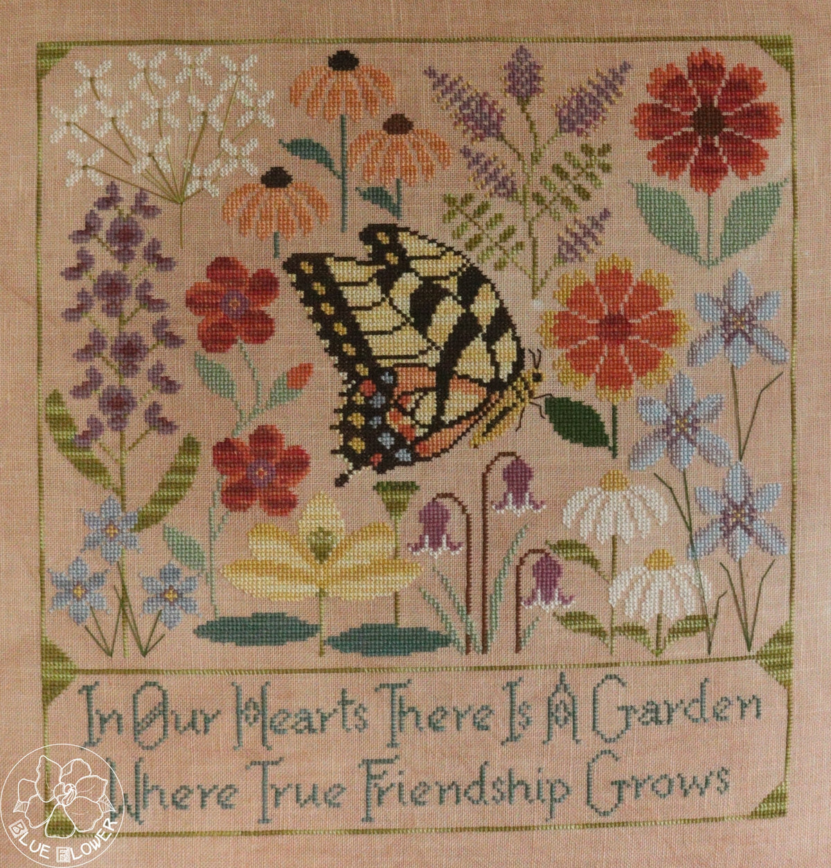 A square cross-stitch sampler featuring a yellow and black swallowtail butterfly surrounded by colorful wildflowers on a beige fabric background, with the embroidered text &
