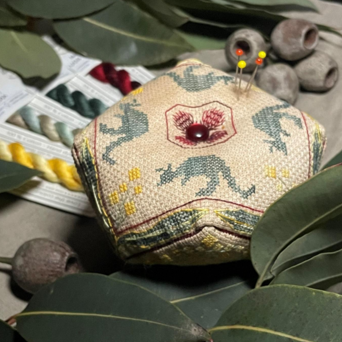cross stitch chart with a kangaroo pattern, colorful threads, and a red embroidered biscorn-roo design on a fabric with a leafy border.