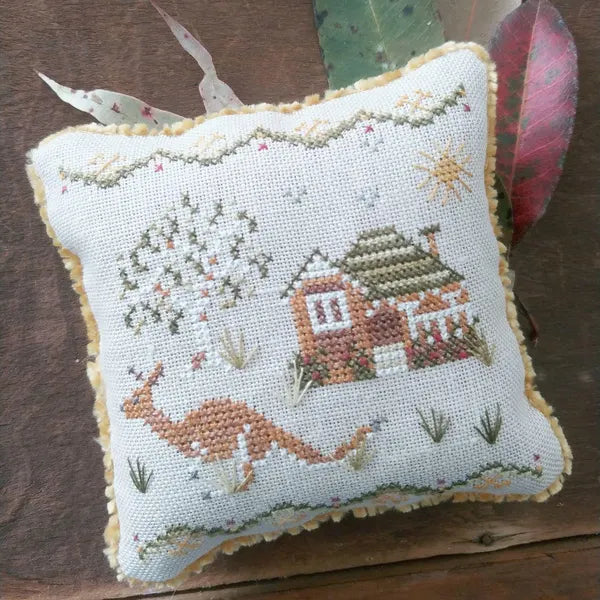 Decorative pillow with cross-stitched design of a federation style house and kangaroo on a wooden surface.