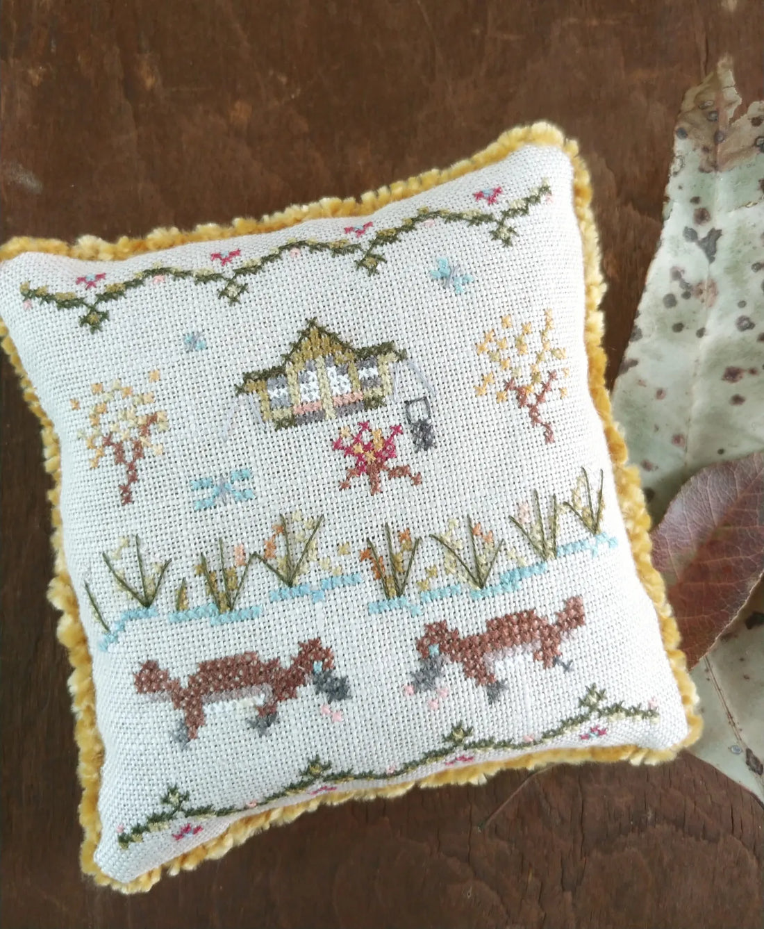 Decorative pillow with cross-stitched design featuring animals and a quiet campsite by a creek on a wooden surface.