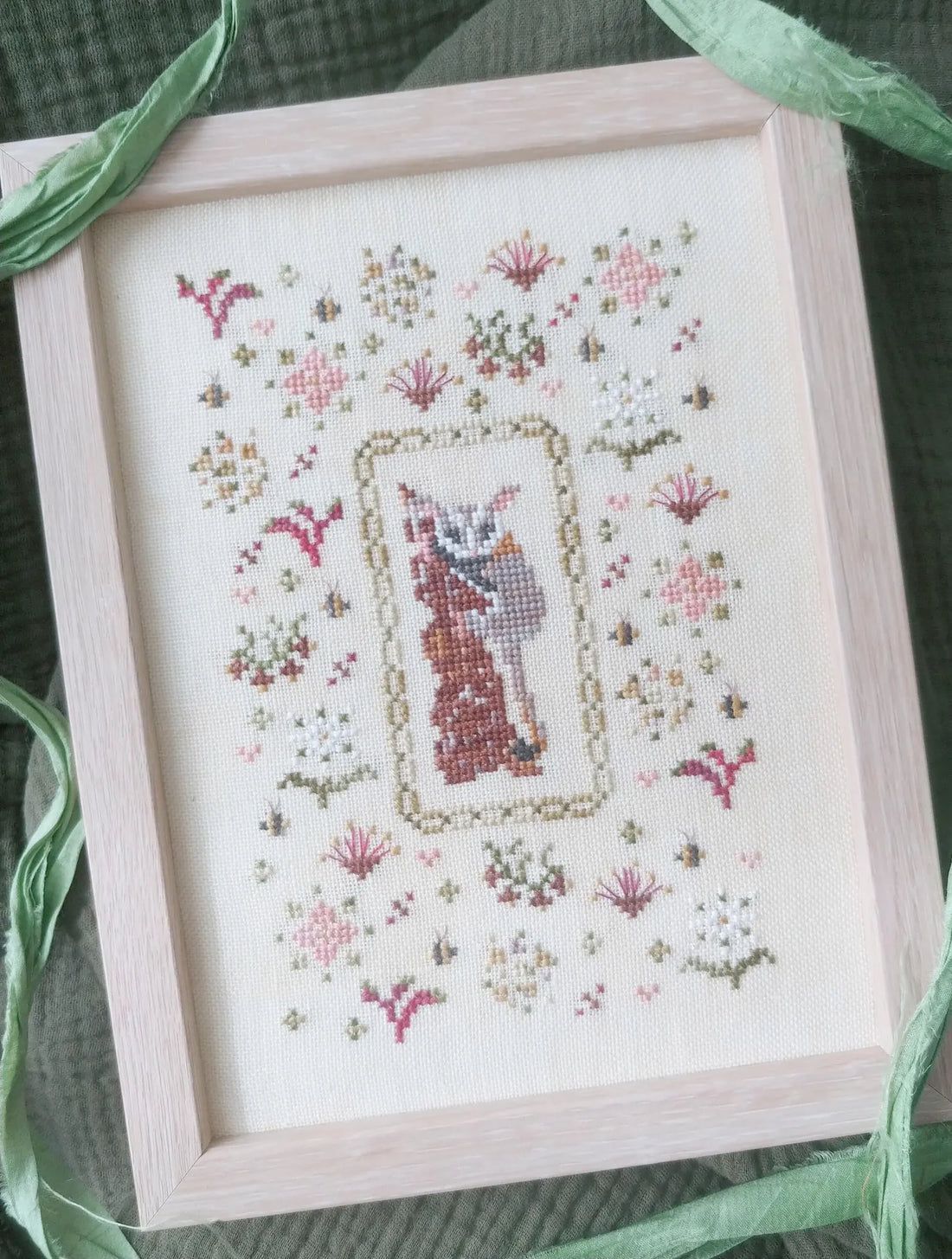Completed cross-stitch embroidery of a cat in a floral frame, framed in a light wood box.