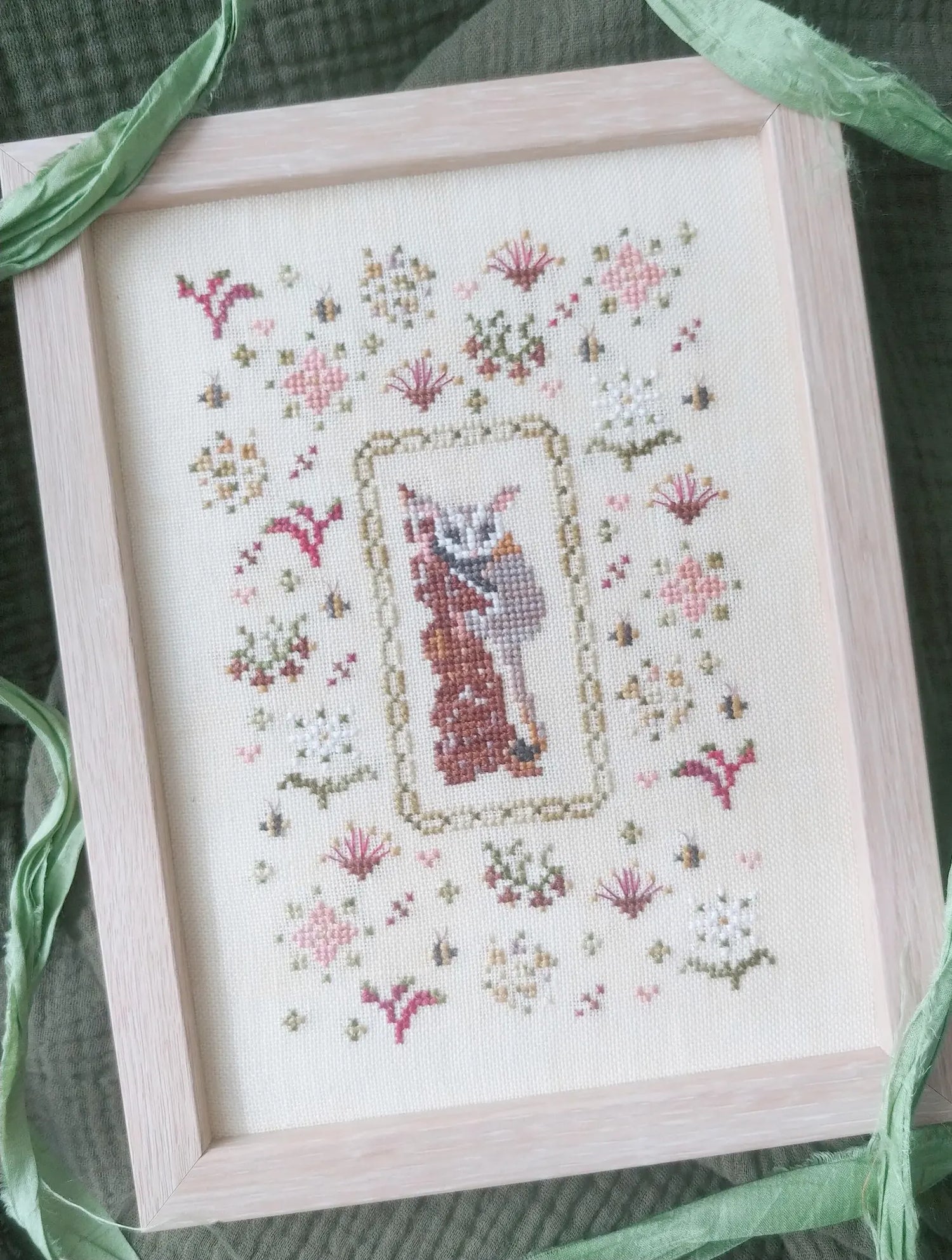 Completed cross-stitch embroidery of a cat in a floral frame, framed in a light wood box.