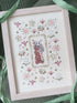 Completed cross-stitch embroidery of a cat in a floral frame, framed in a light wood box.