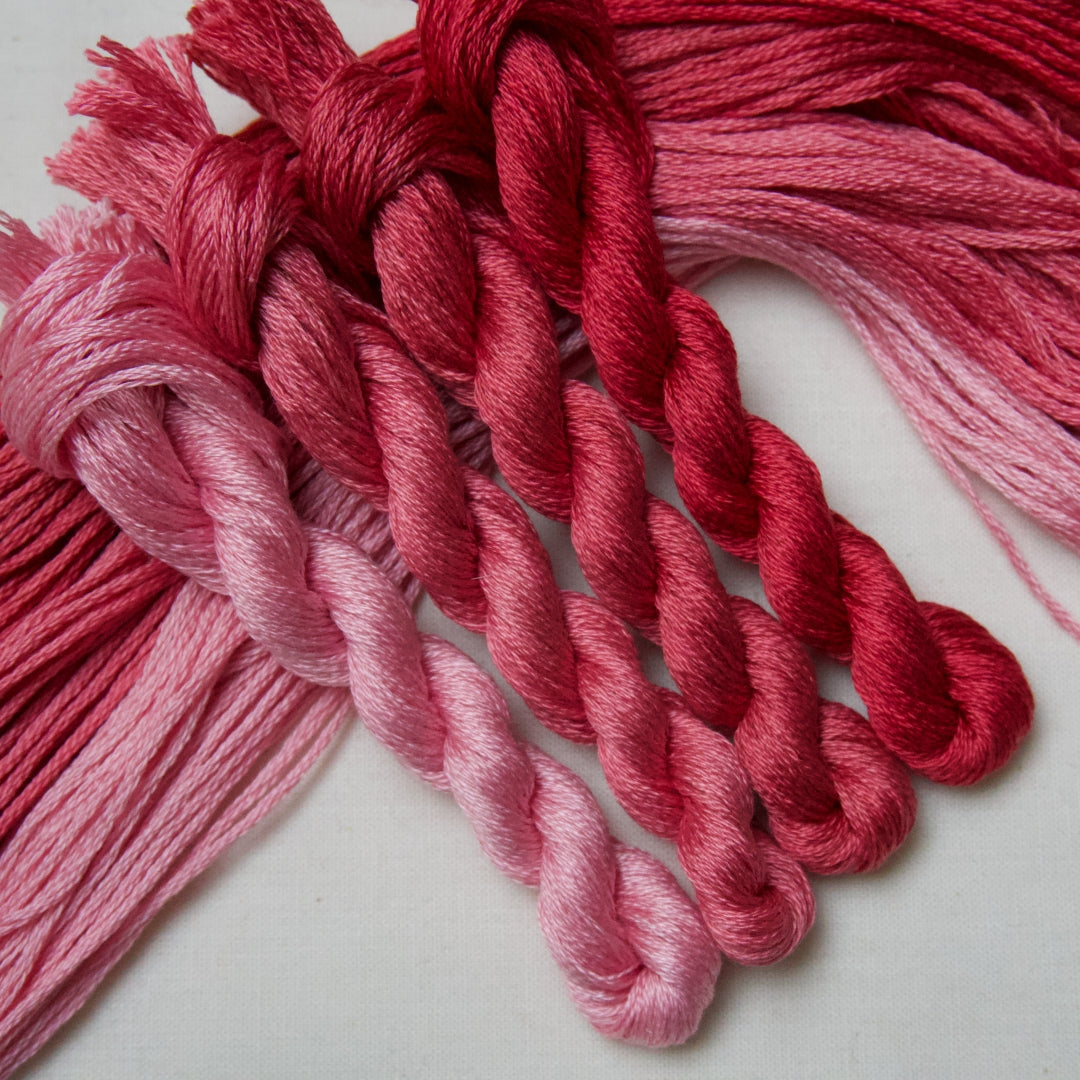 Four skeins of embroidery floss in a vertical gradient of shades from light pastel pink to deep crimson red, arranged on a light gray background.