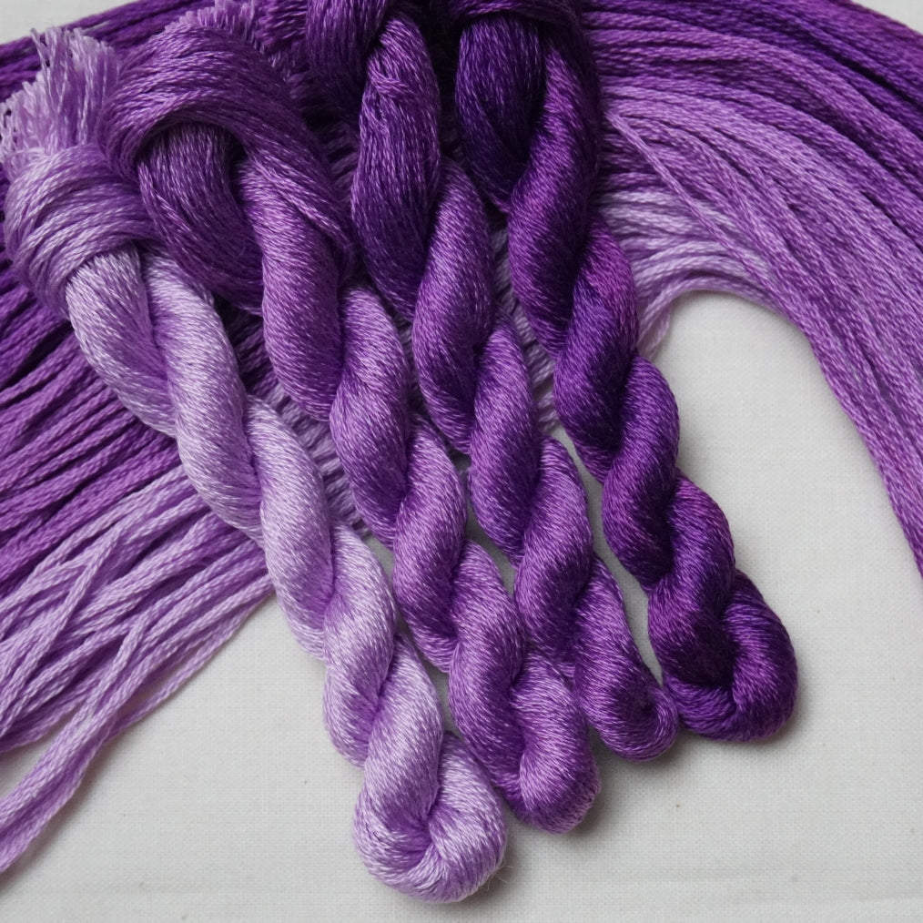Four twisted skeins of yarn in shades of purple, arranged on a plain light background.