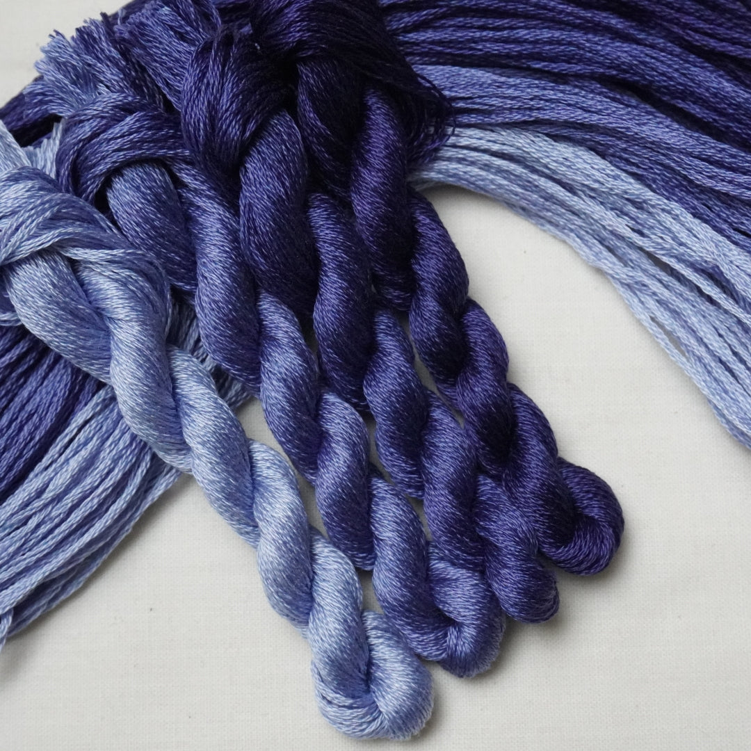 Four braided skeins of yarn in a blue-purple ombre gradient, arranged on a light off-white background.