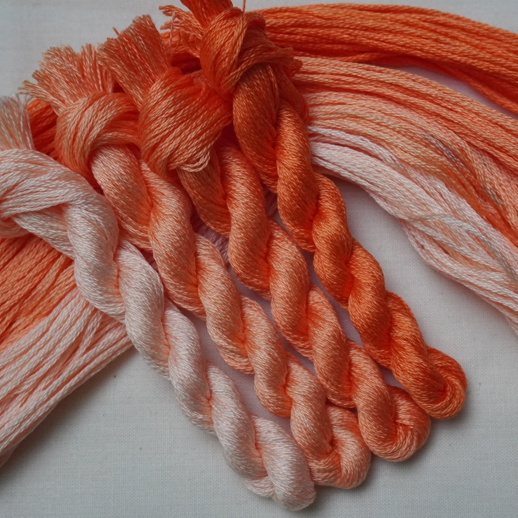 A close-up of four twisted embroidery threads in a peach, orange, and pale peach gradient, arranged on a light neutral fabric background.